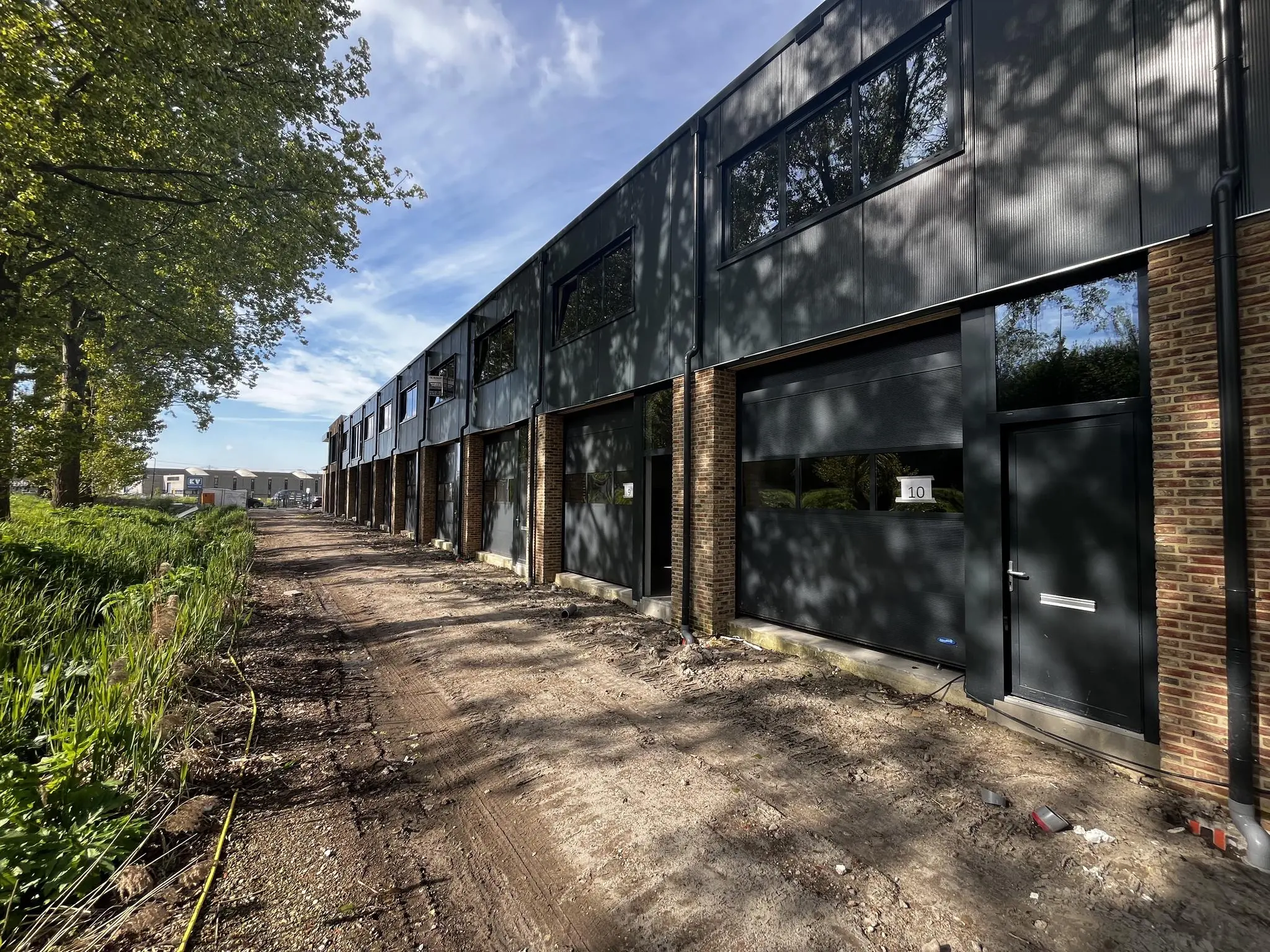 Nieuwbouw bedrijfsunits aan de Broekdijk West 19-10, gelegen langs een onverharde weg met bomen en groenstroken.